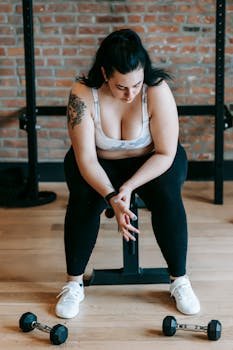 Full body of obese tired female in activewear having break in training with dumbbells in gym
